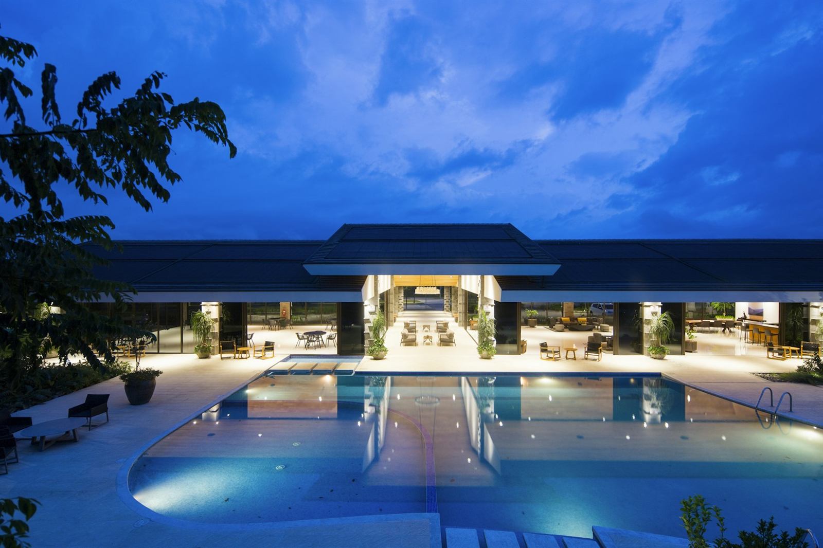 A modern luxury hotel with a warmly lit lounge and an illuminated infinity pool at blue hour